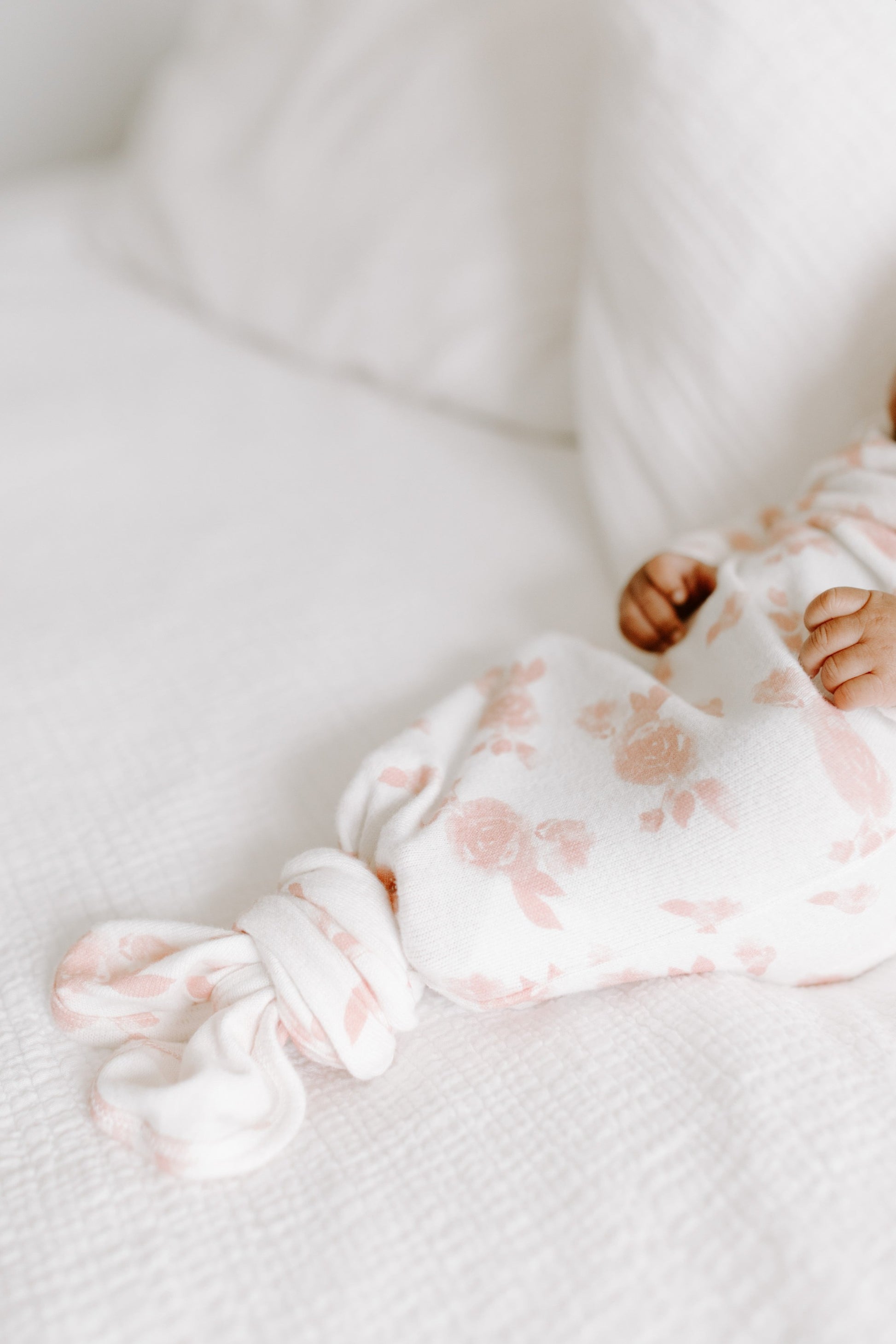 Naujagimis aprengtas kreminės spalvos smėlinuku su rausvomis rožytėmis gulintis ant lovos su balta patalyne