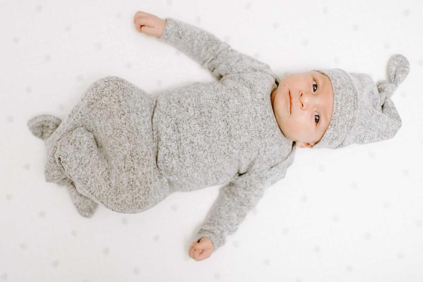 Ant lovos su baltos spalvos antklode ir pilkais taškučiais gulintis kūdikis aprengtas pilku smėlinuku ir kepurėle