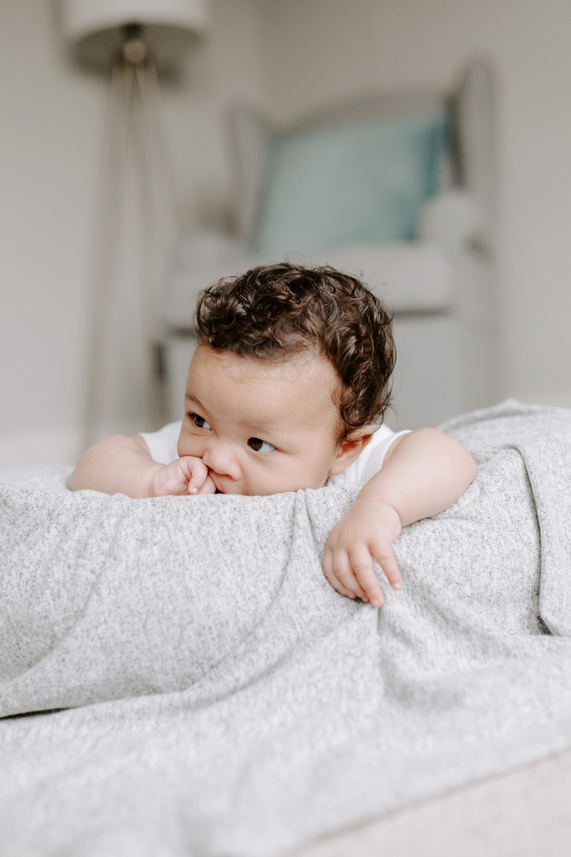 Žiūrintis į šoną kūdikis gulintis kambaryje ant lovos su pilkos spalvos antklode. Už lovos stovi krėslas ir šviestuvas