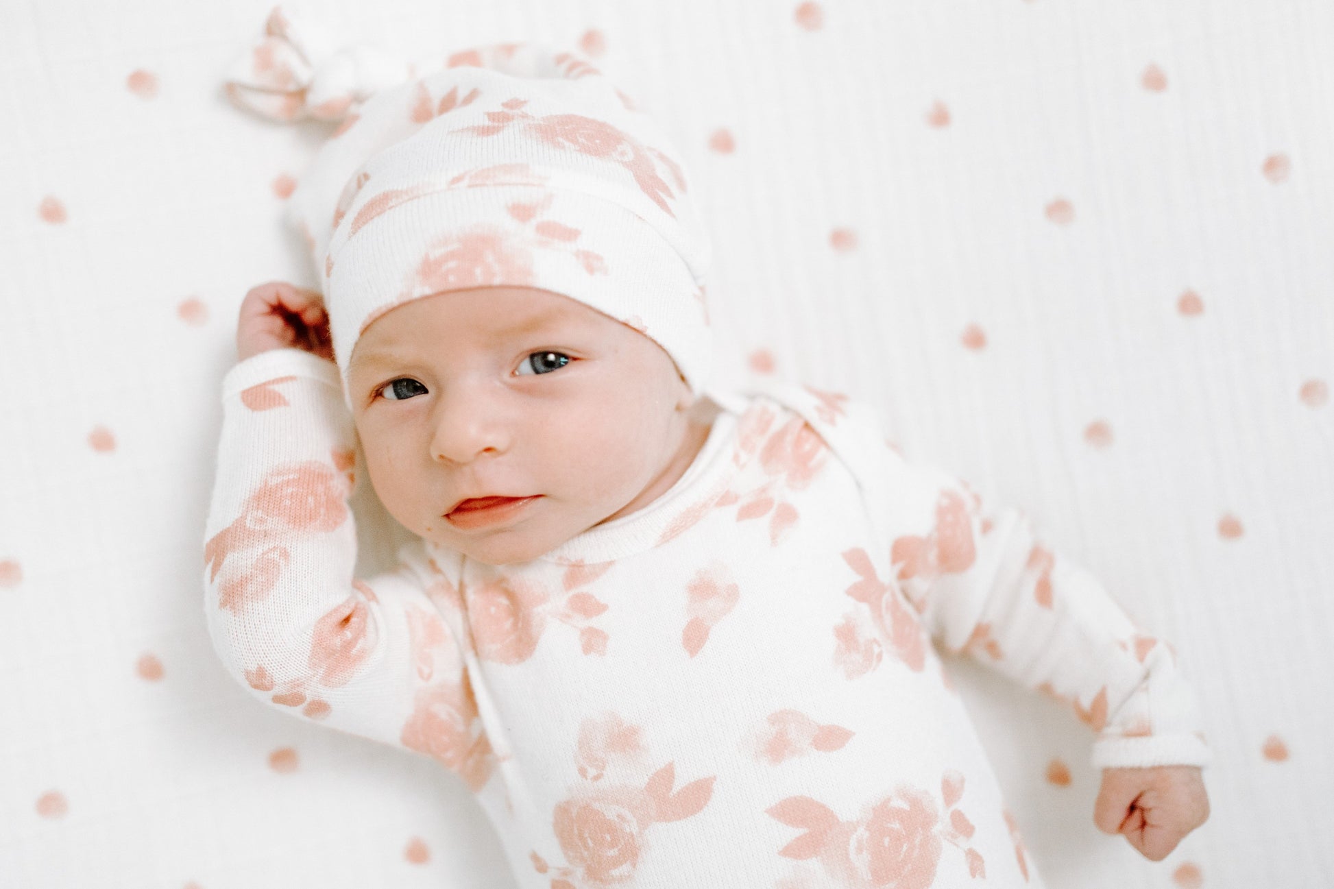 Žiūrintis į objektyvą kūdikis su kreminės spalvos smėlinuku ir kepuryte ant to pačio rašto antklodės su rausvomis rožytėmis