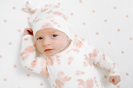 Žiūrintis į objektyvą kūdikis su kreminės spalvos smėlinuku ir kepuryte ant to pačio rašto antklodės su rausvomis rožytėmis