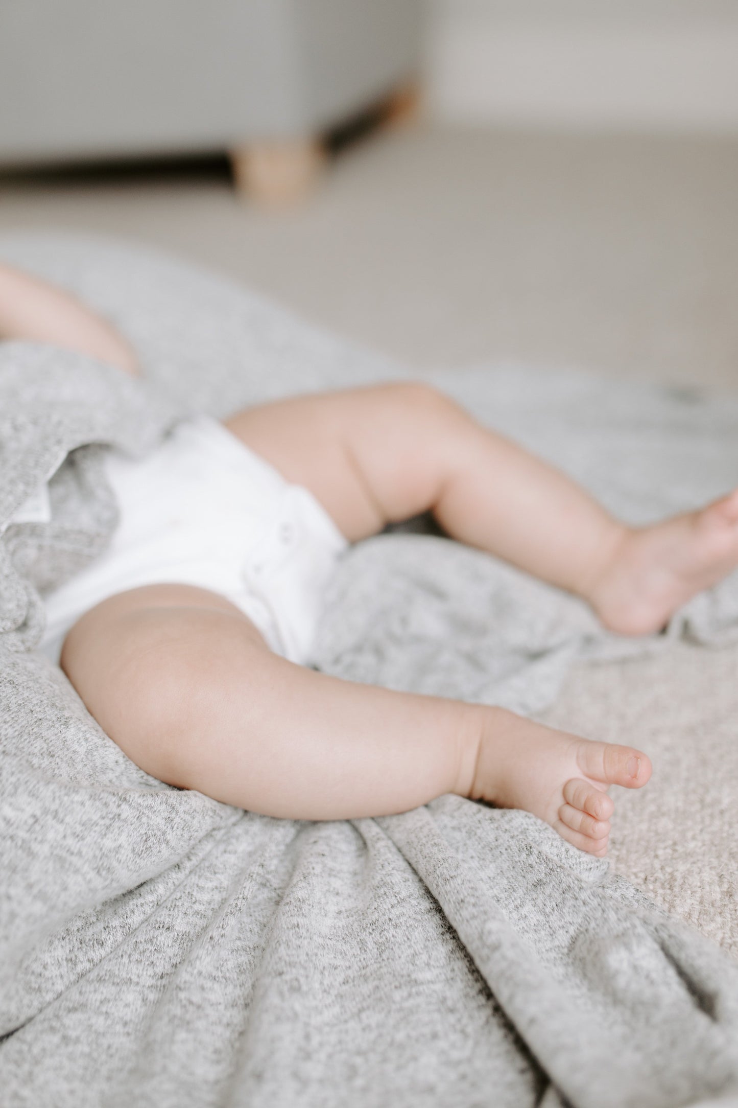Ant lovos su pilkos spalvos antklode gulintis kūdikis nuogomis kojytėmis aprengtas baltu smėlinuku. Fone matosi sofos kampas