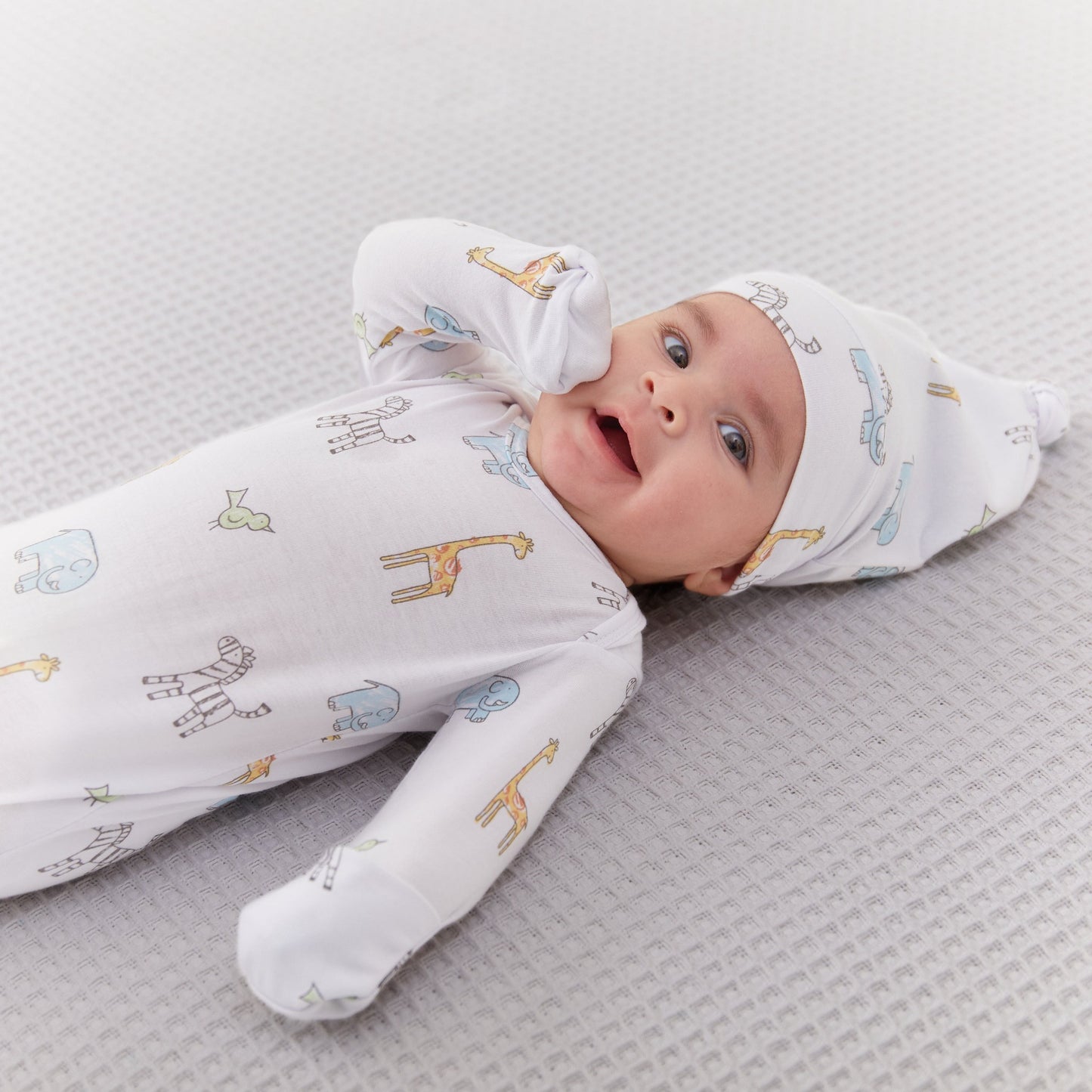 Kūdikis aprengtas balto smėlinuko ir kepurėlės komplektu su gyvūnėliais: žirafomis, drambliukais, zebriukais ir paukščiukais 