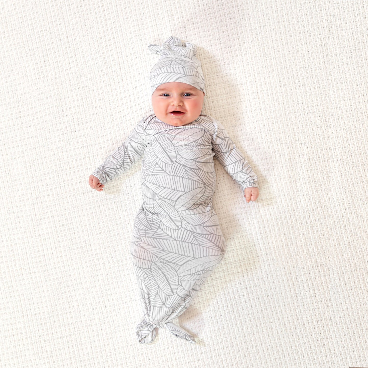 Išsišiepęs, laimingas kūdikis su smėlinuko ir kepurėlės komplektu su baltai pilkais lapeliais ant kreminės spalvos kilimo