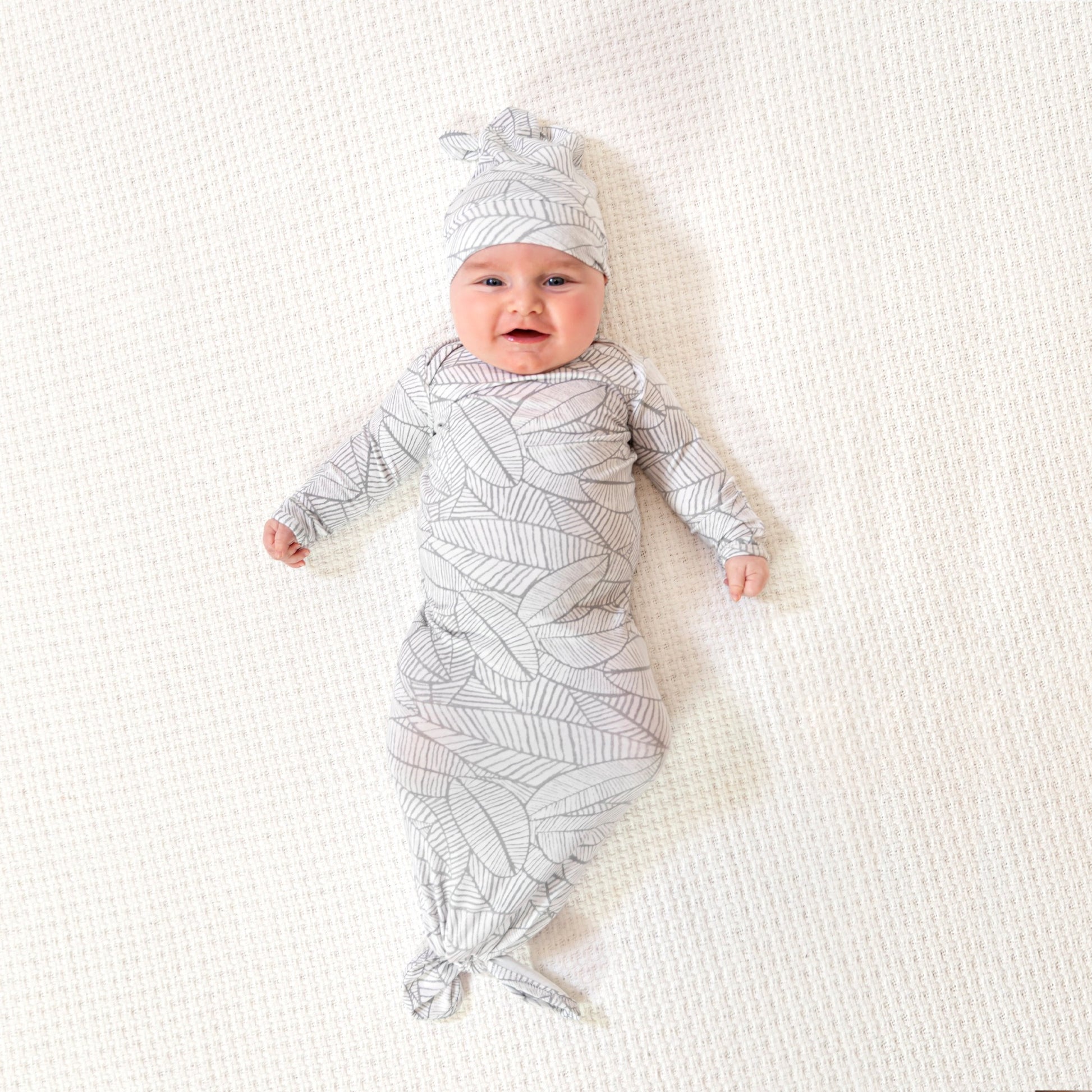 Išsišiepęs, laimingas kūdikis su smėlinuko ir kepurėlės komplektu su baltai pilkais lapeliais ant kreminės spalvos kilimo
