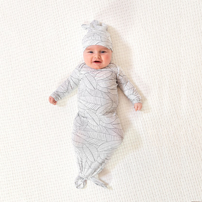 Išsišiepęs, laimingas kūdikis su smėlinuko ir kepurėlės komplektu su baltai pilkais lapeliais ant kreminės spalvos kilimo