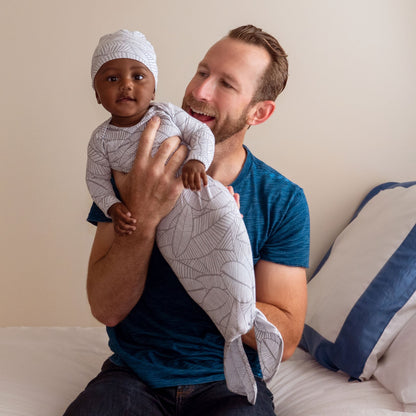 Besišypsantis vyras ant rankų laiko susidomėjusį kūdikį su smėlinuko ir kepurėlės komplektu su baltai pilkais lapeliais