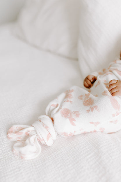 Naujagimis aprengtas kreminės spalvos smėlinuku su rausvomis rožytėmis gulintis ant lovos su balta patalyne 