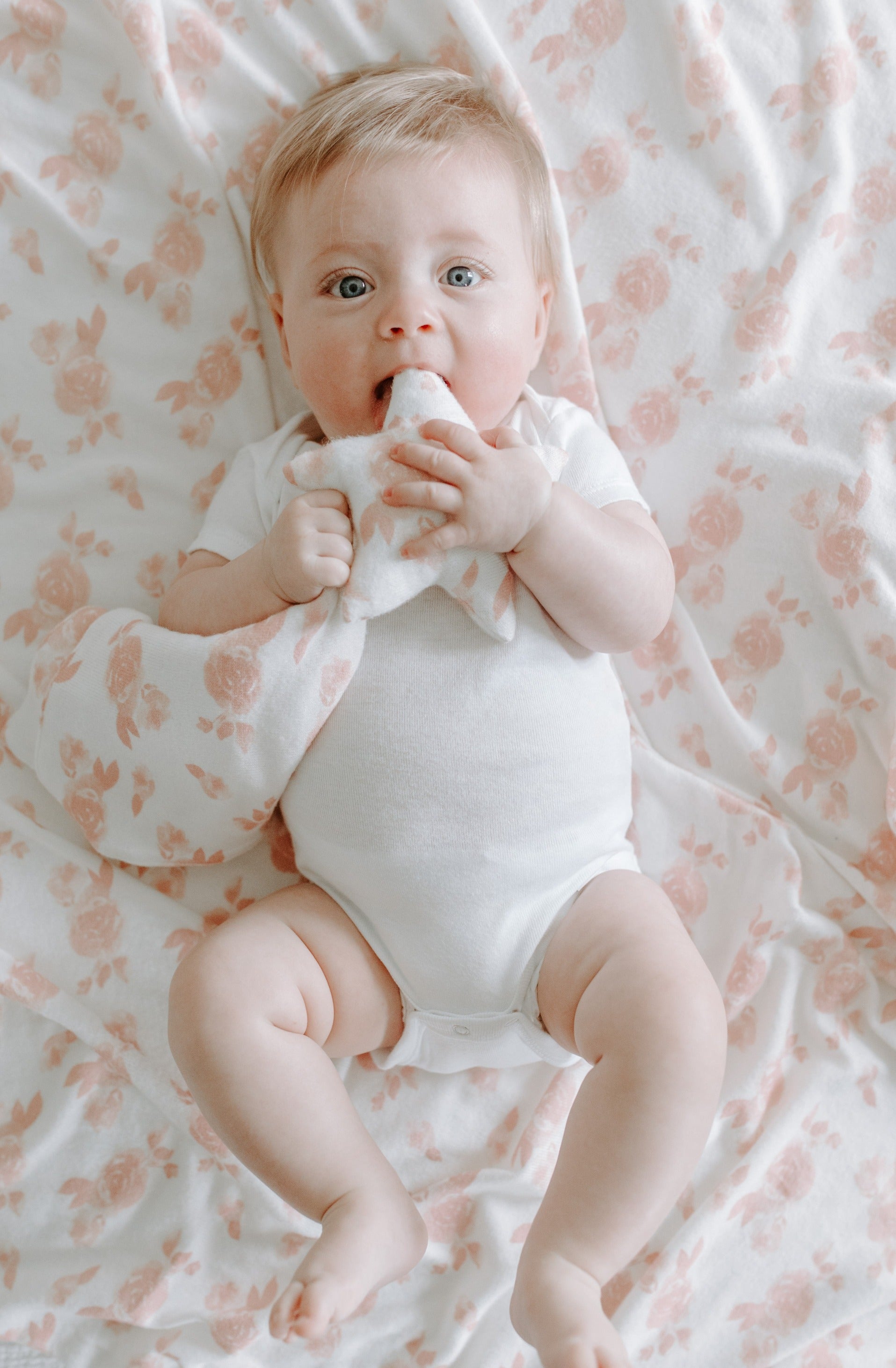 Žiūrintis į objektyvą linksmas kūdikis su smėlinuku, gulintis ant kreminės spalvos antklodės su rausvomis rožytėmis